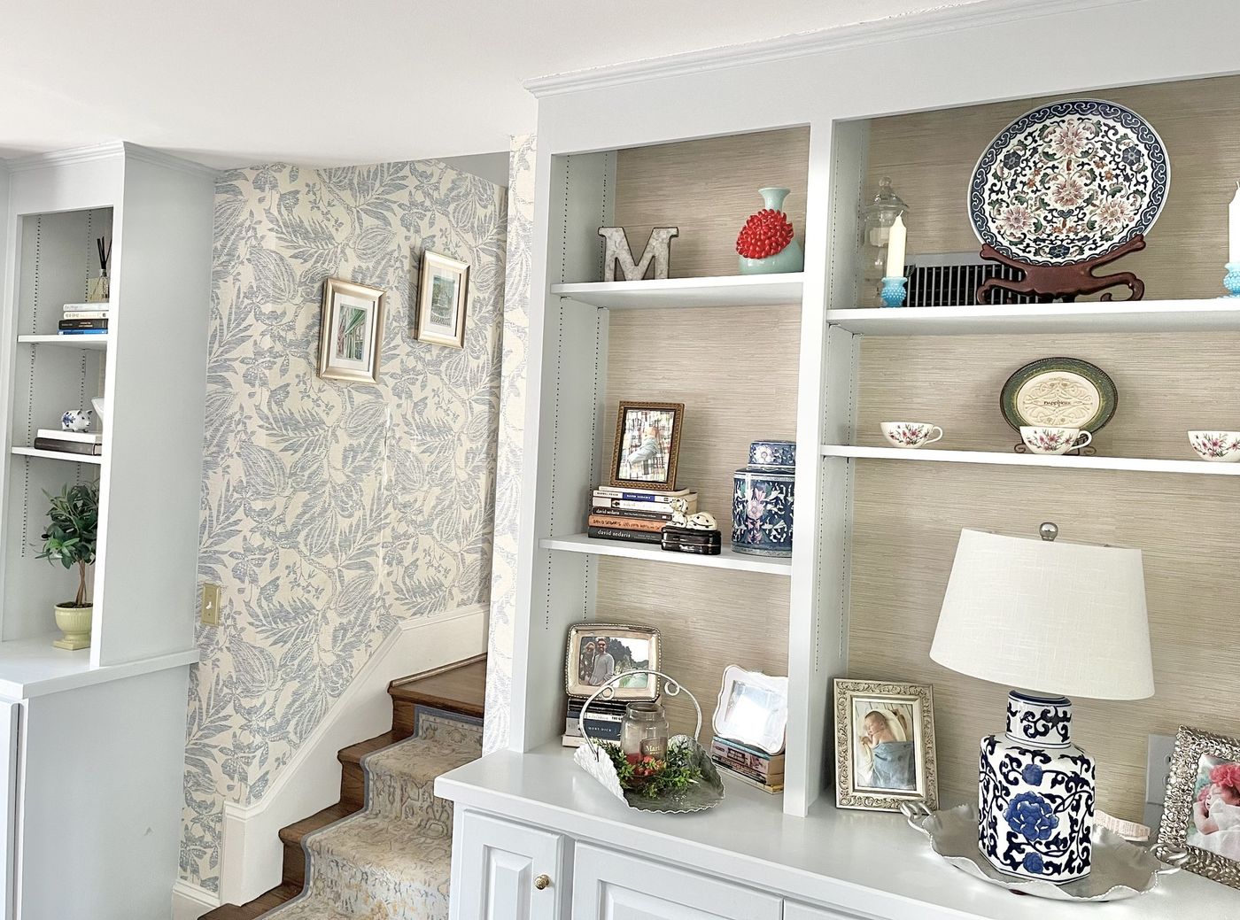 Built-in shelves beside a stair hall wrapped in blue botanical wallpaper