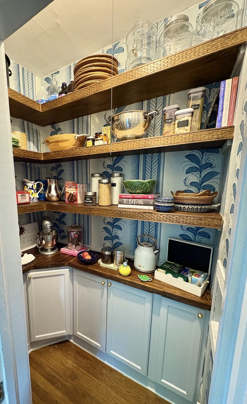 Pantry with leaf-print wallpaper, rattan-edged wood shelves, and grey lower cabinets