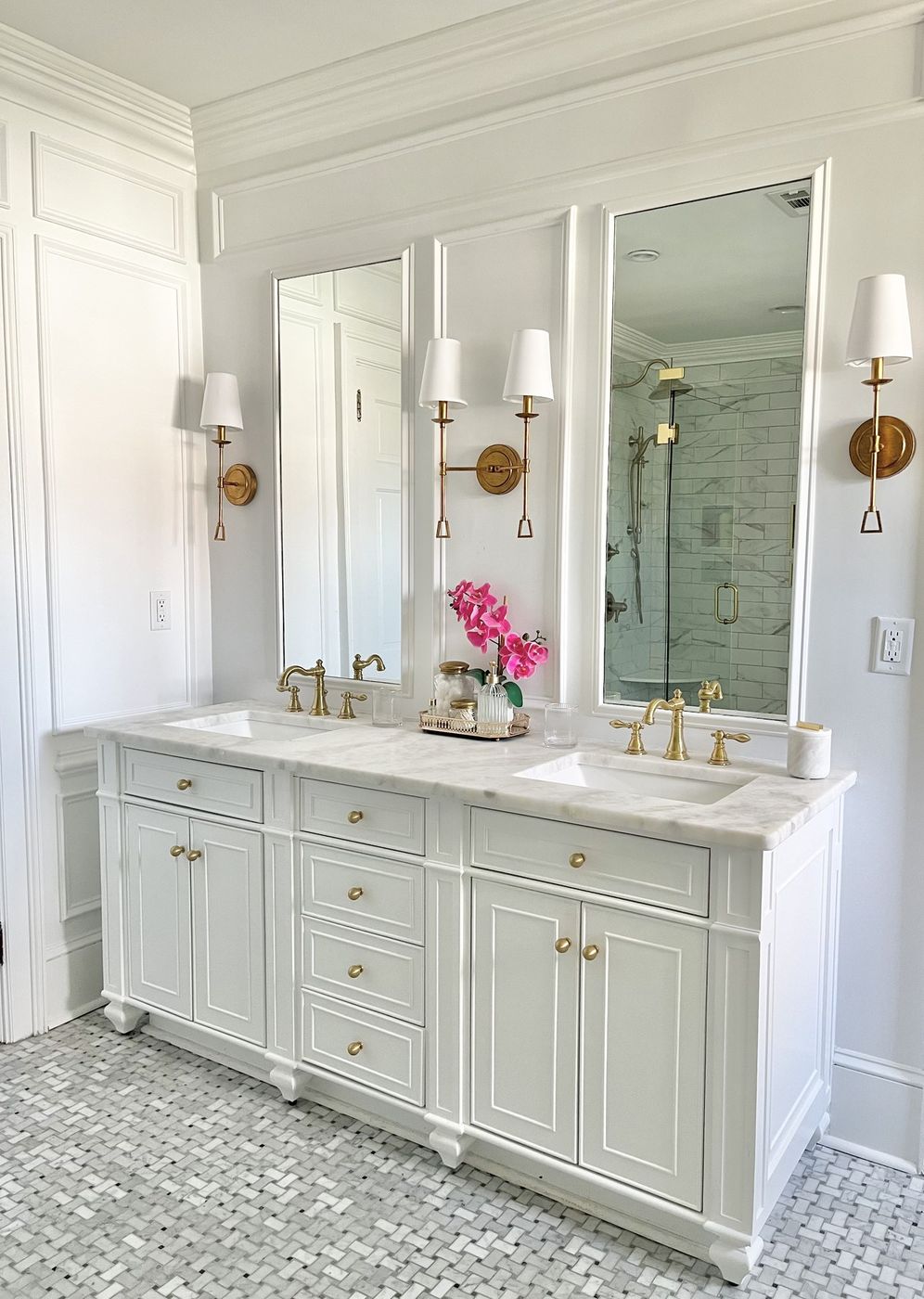 Primary bath with a white double vanity, brass sconces, and basketweave marble floor